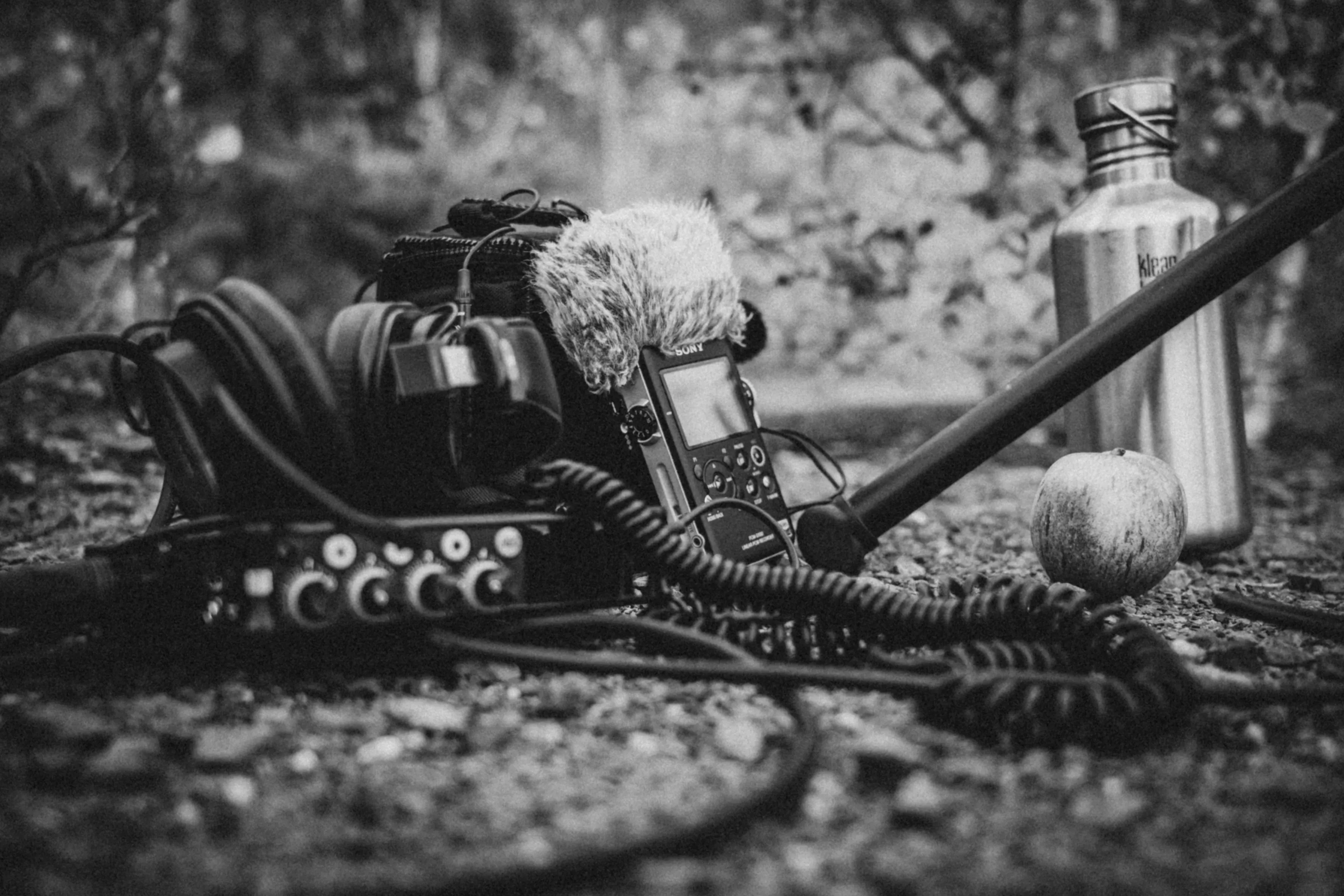 Audio-Equipment auf kiesigem Untergrund: Recorder mit Fell-Windschutz, Kopfhörer, kleiner Mischer, Tonangel — schwarzweiß.