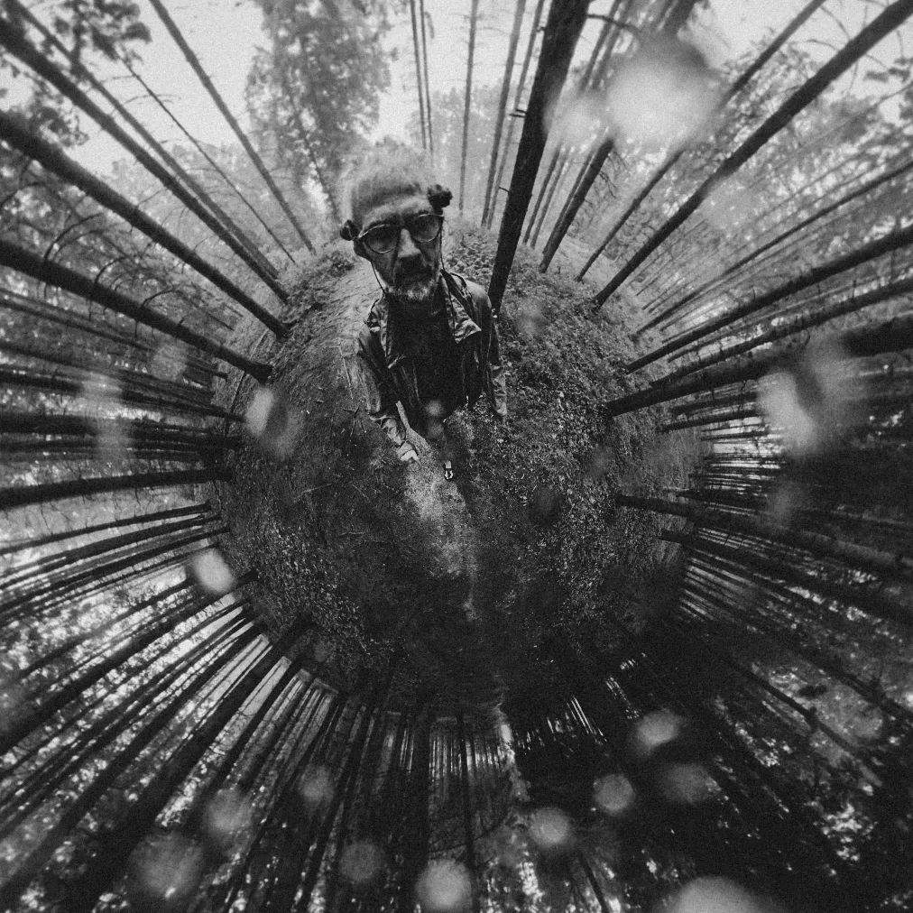 Tiny-Planet-Verzerrung eines Waldes mit einer Person in der Mitte, die binaurale Mikrofone an den Ohren trägt, schwarzweiß.