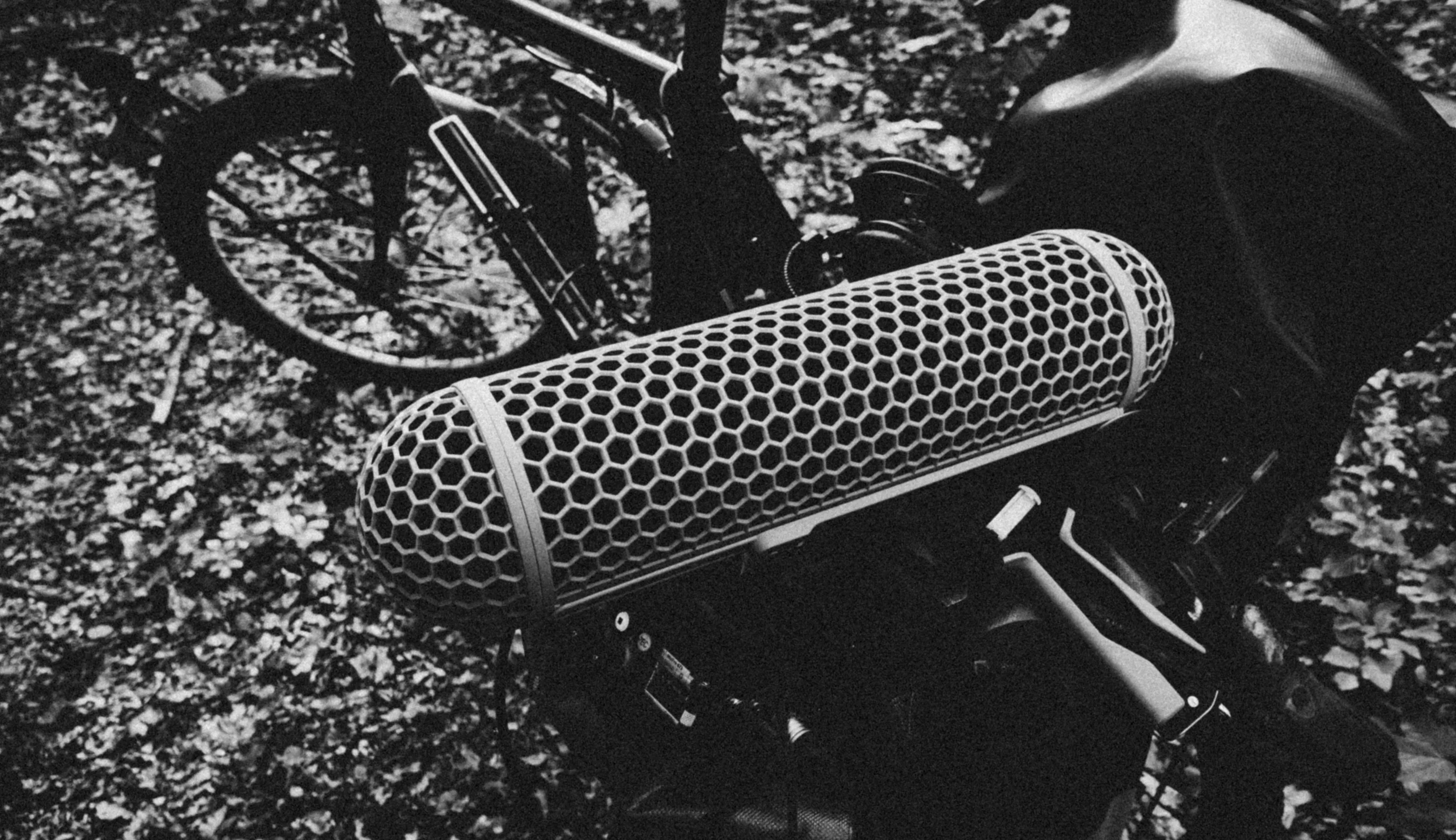 Nahaufnahme eines zylindrischen Mikrofon-Windschutzes (Blimp) vor einem im Waldboden geparkten Fahrrad, schwarzweiß.
